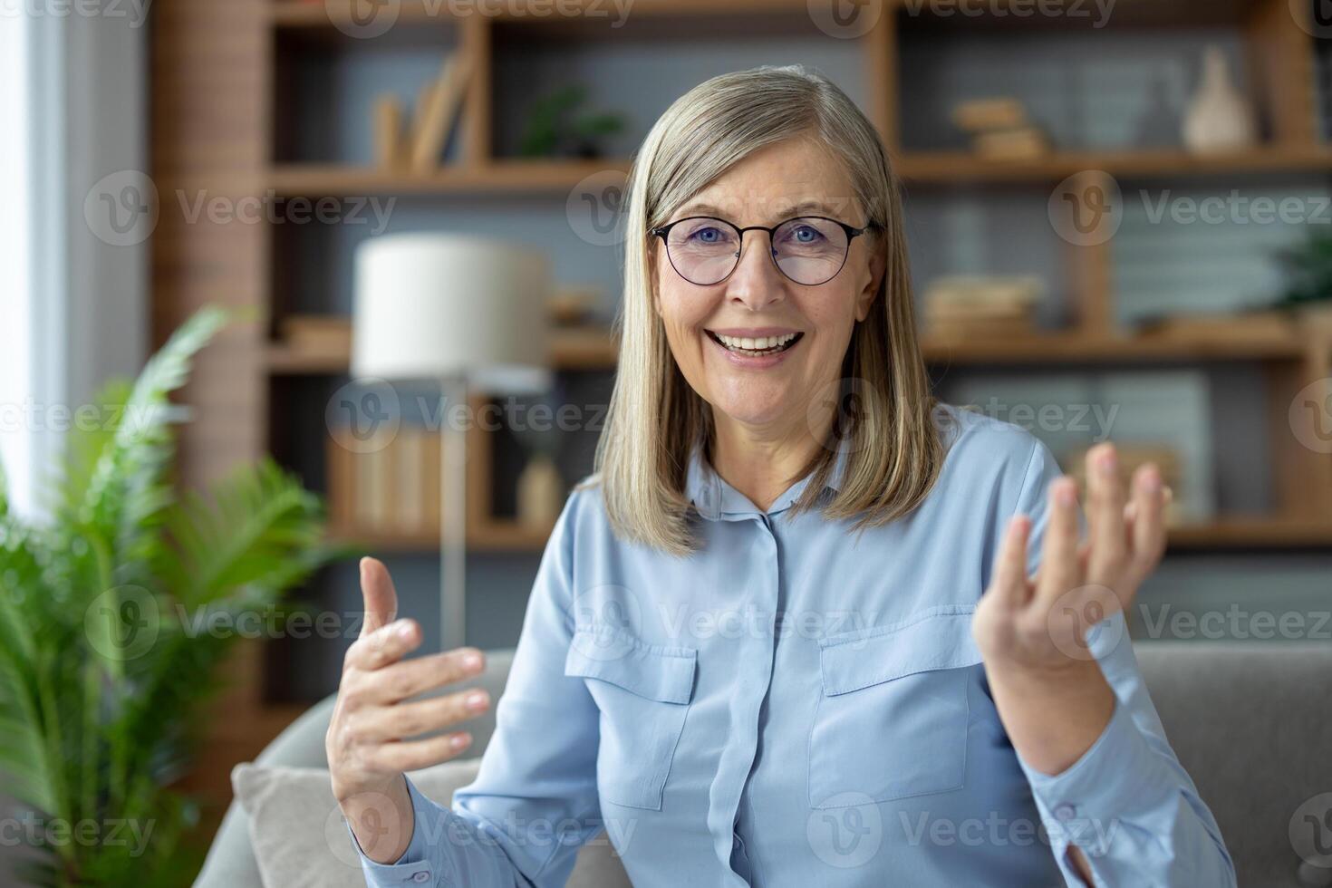Senior woman with glasses engaging in a call while looking and talking to the camera. She seated in a cozy living room with shelves and plants in the background, smiling warmly and gesturing. photo