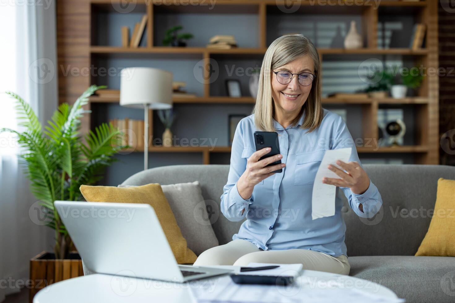 Senior woman sitting on couch using smartphone and computer while checking receipts. Concept of remote work, home finance management, and online accounting. Modern cozy home interior. photo