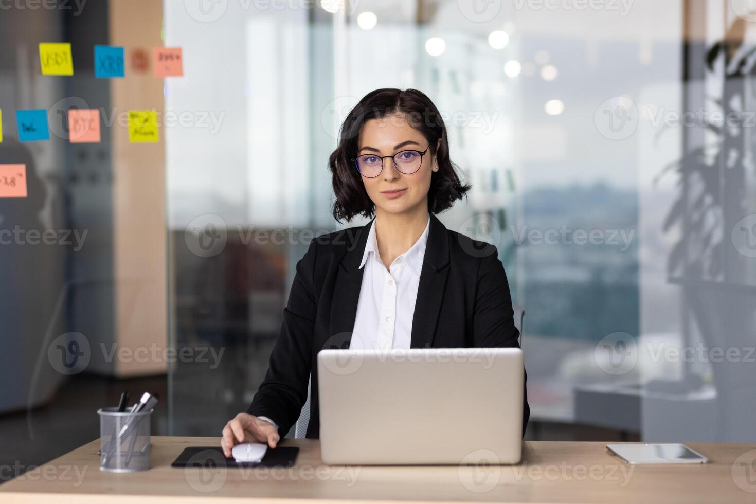 Caucasian woman in eyewear with short haircut sitting by desktop and holding hand on computer mouse in glassy wall office. Perspective company employee starting new day in personal workspace. photo