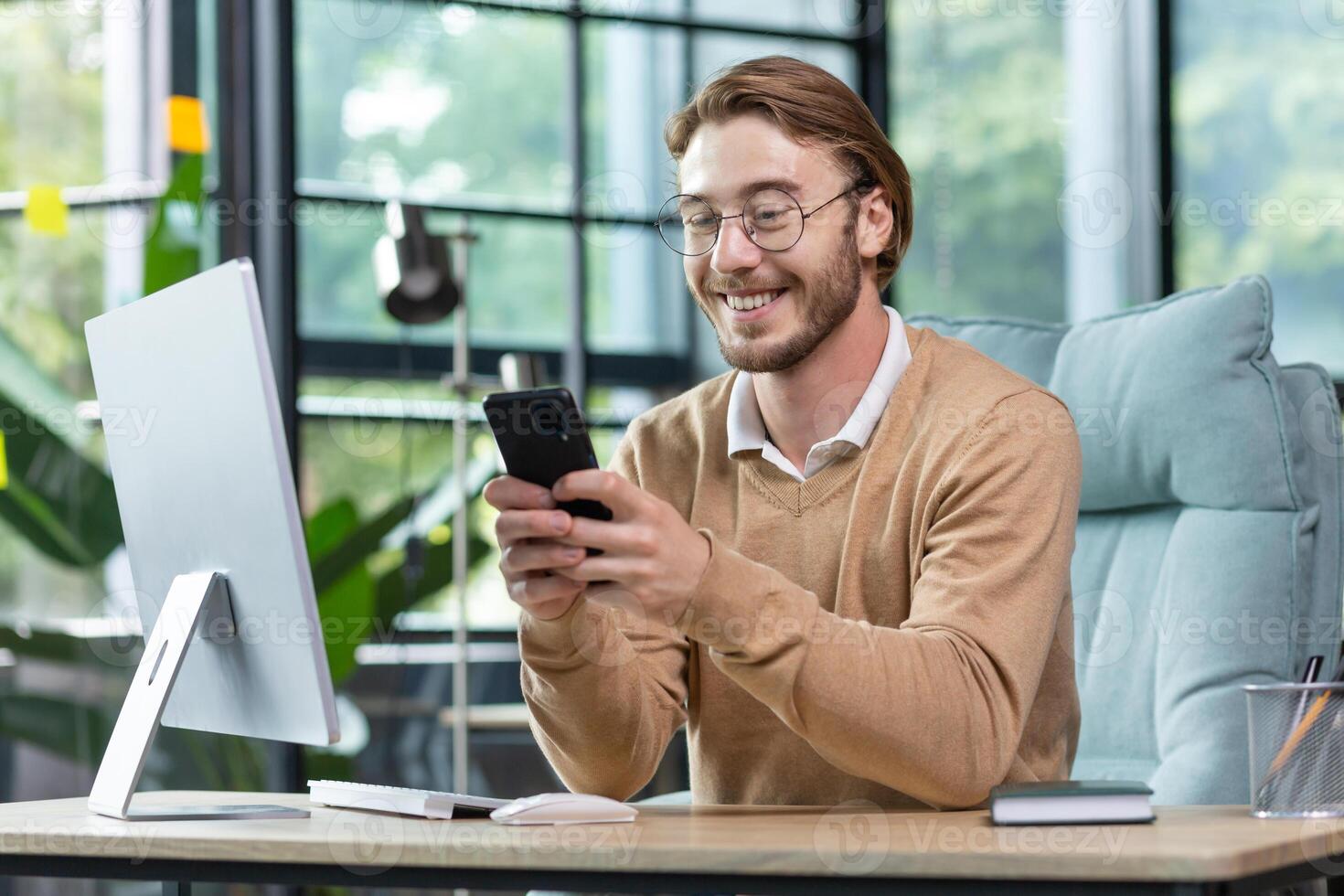 Young handsome man businessman, freelancer sits in the office at the computer and uses the phone. Type messages, chstting,, read news, smile. photo