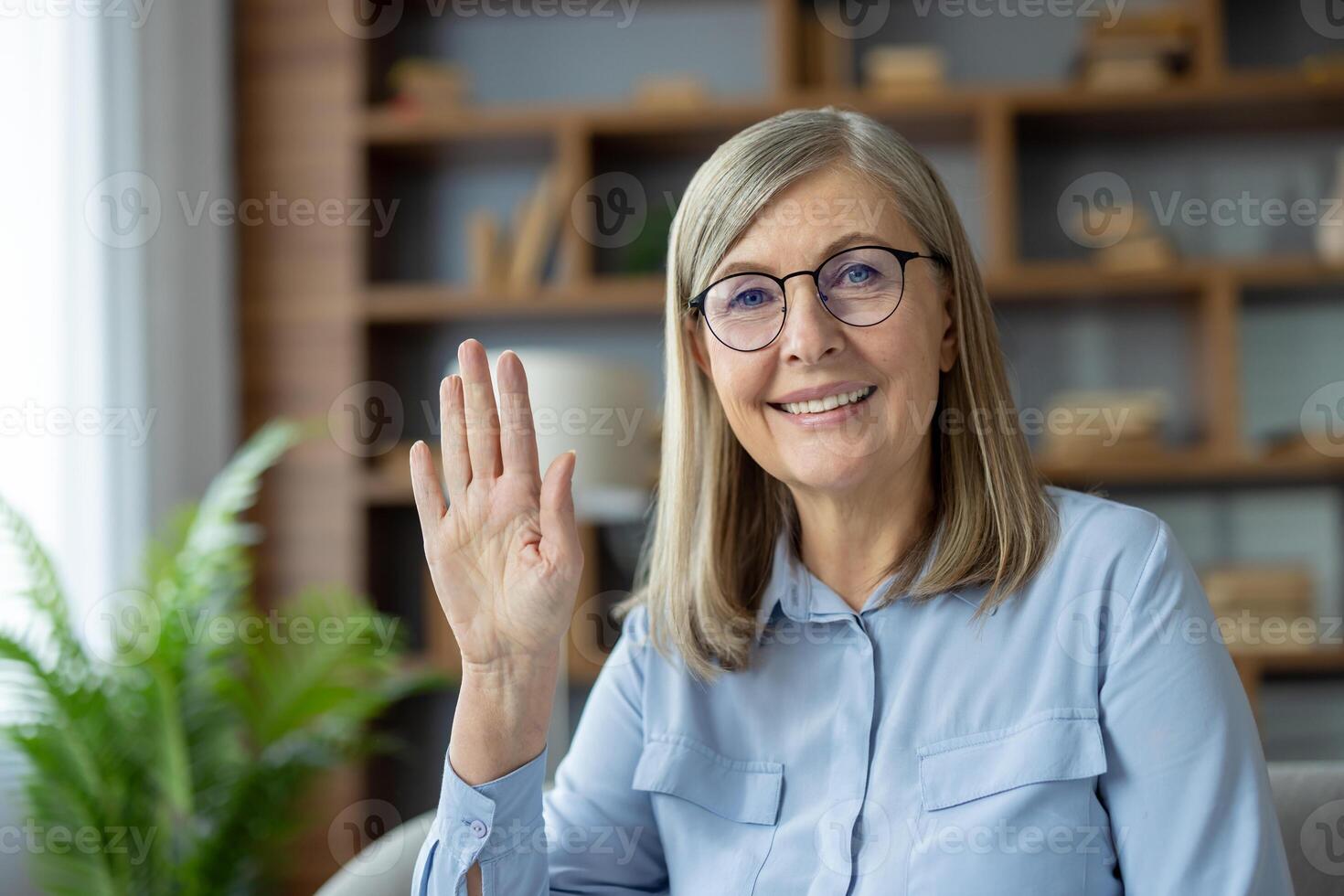 Senior woman with glasses smiling and waving while engaging in a call from home, representing communication and connection. photo
