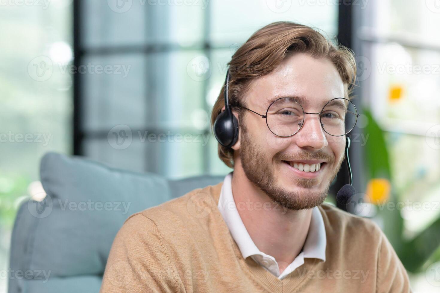 Portrait of young man with headset for call inside office, close-up tech support worker in glasses smiling and looking at camera, businessman blond customer service online help. photo