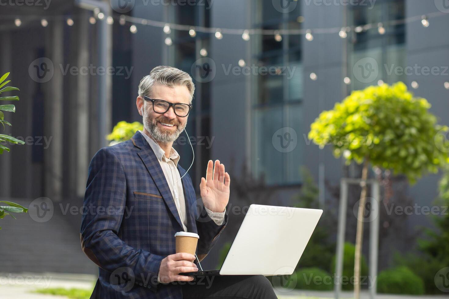 Friendly senior executive in business attire waving and working on a laptop outside a modern office building with a coffee cup. photo