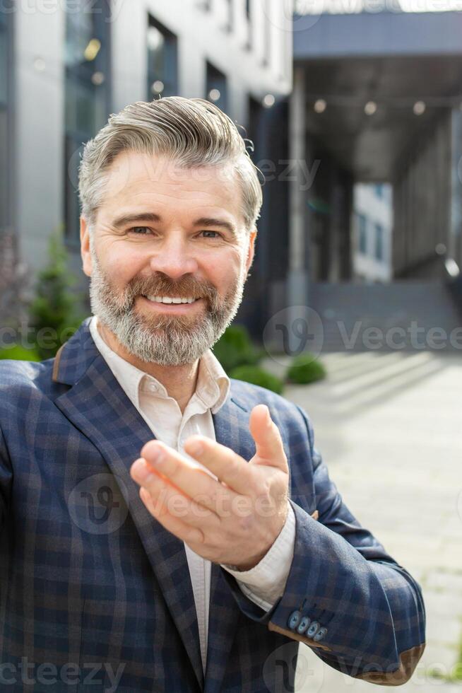 Cheerful experienced senior professional offering a handshake in a friendly gesture, standing confidently outside an office. photo
