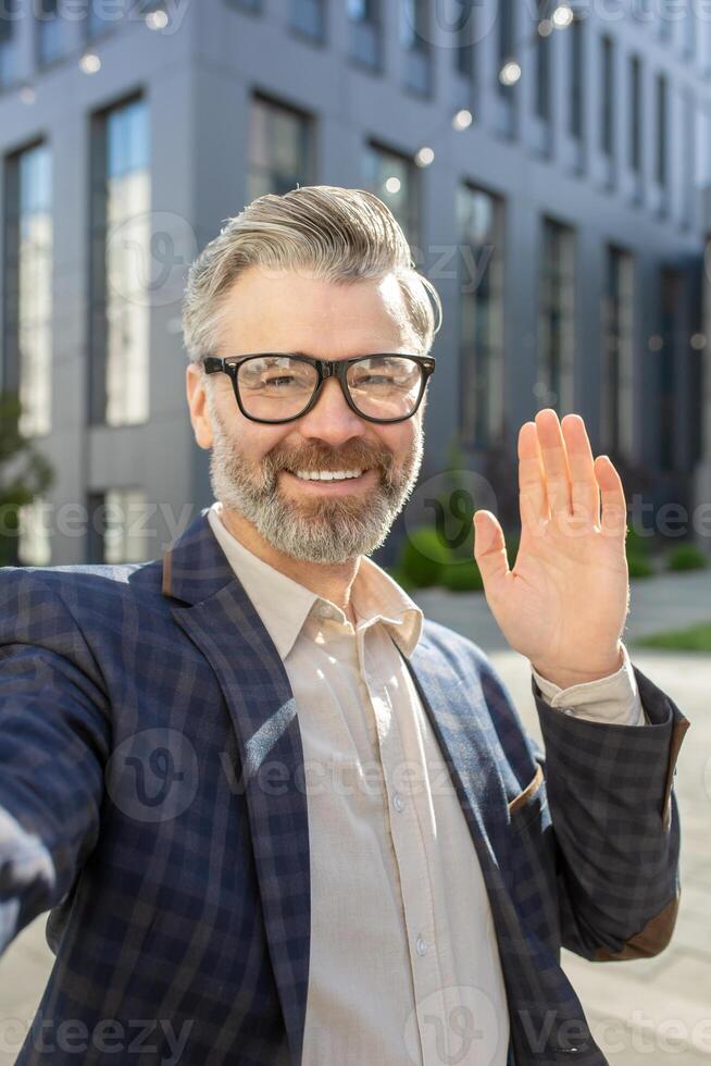 Cheerful senior male executive with glasses greeting with a hand wave, standing outside a modern office building in a suit. photo