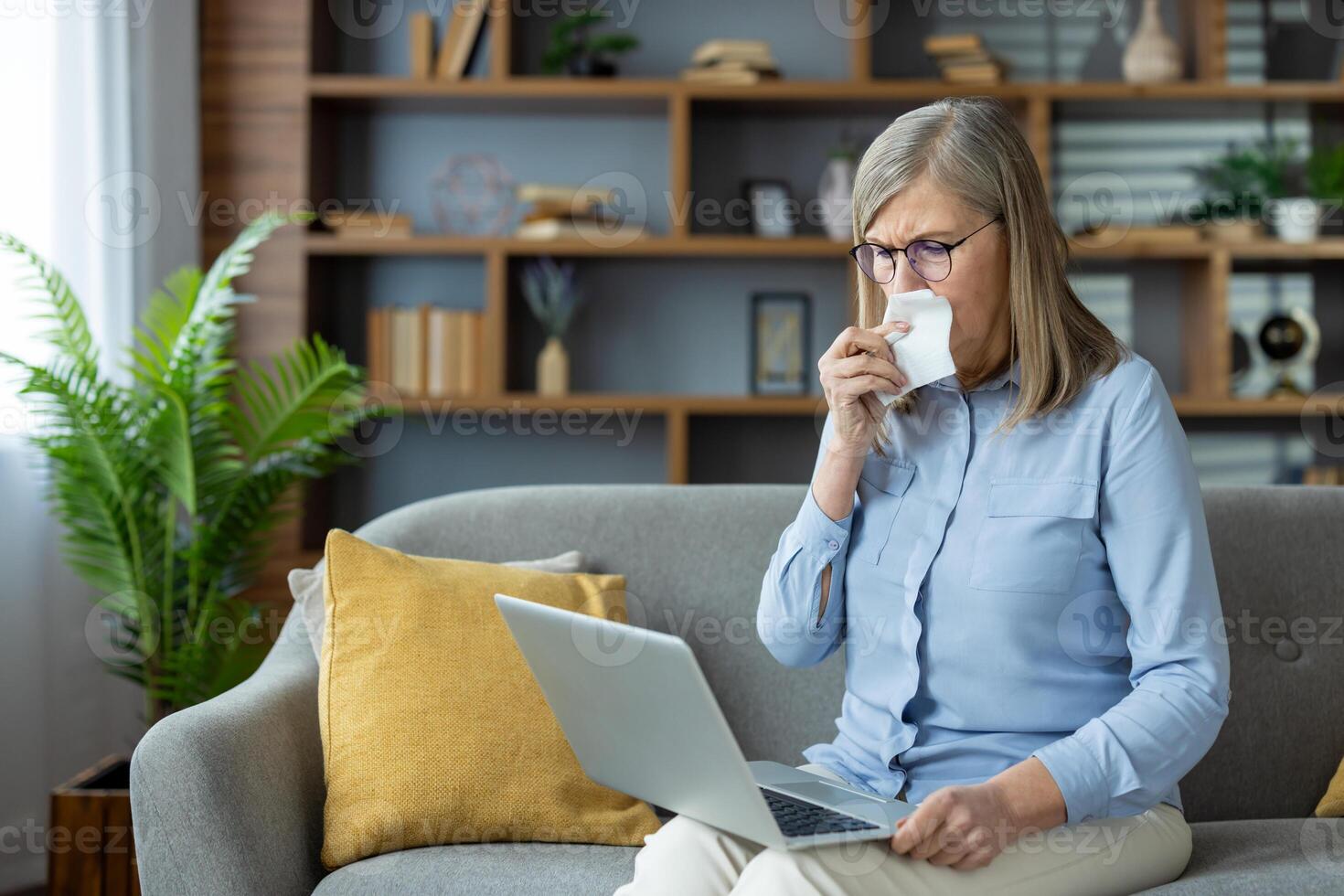 A senior woman crying and using a tissue during an online consultation with a psychologist at home on a laptop. photo