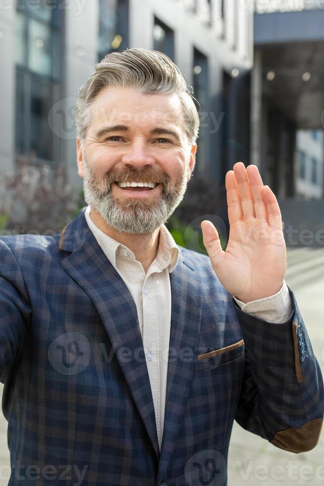 Smiling mature businessman in a stylish suit waving hello, standing outdoors by an office building, exuding leadership. photo