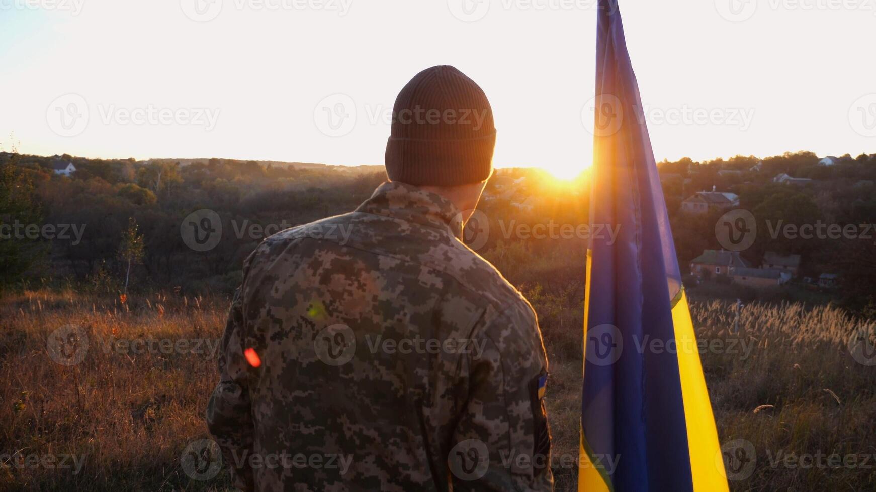 Male ukrainian army soldier stands with national banner against background of beautiful sunset. Young man in camouflage uniform with Ukraine flag as symbol of victory against russian aggression photo