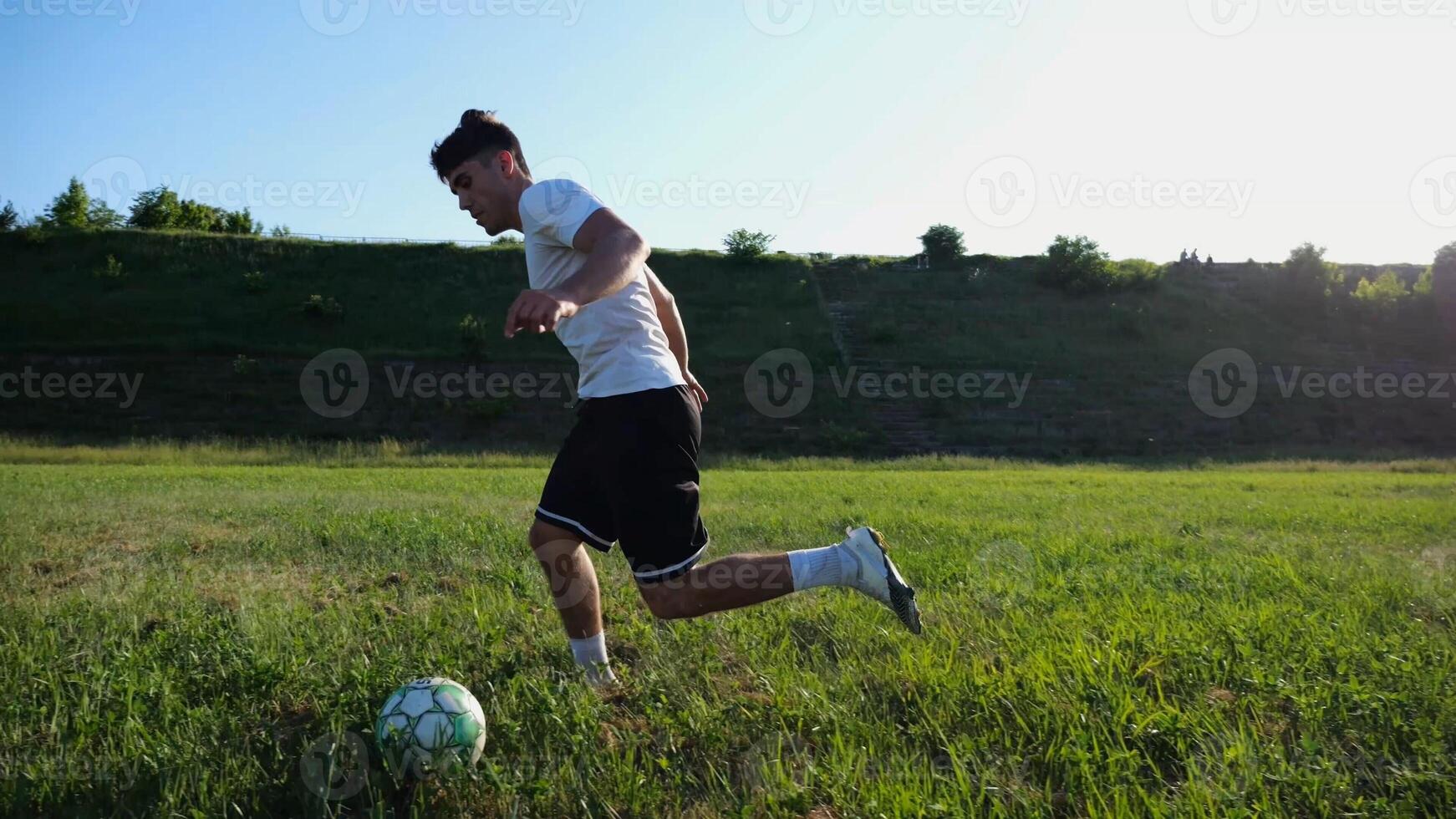 Professional footballer in uniform shows tricks with ball while running at green field. Sportsman kicks ball during jogging at meadow on sunny day. Young man training at stadium. Freestyle football photo