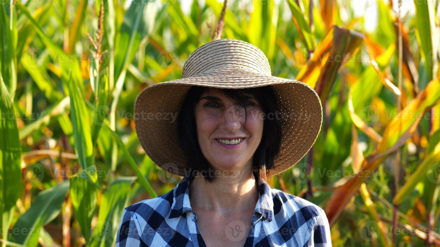 Happy smiling female farmer looks into camera standing near corn field. Portrait of adult beautiful agronomist in straw hat with maize meadow at background. Agricultural business concept. Close up photo