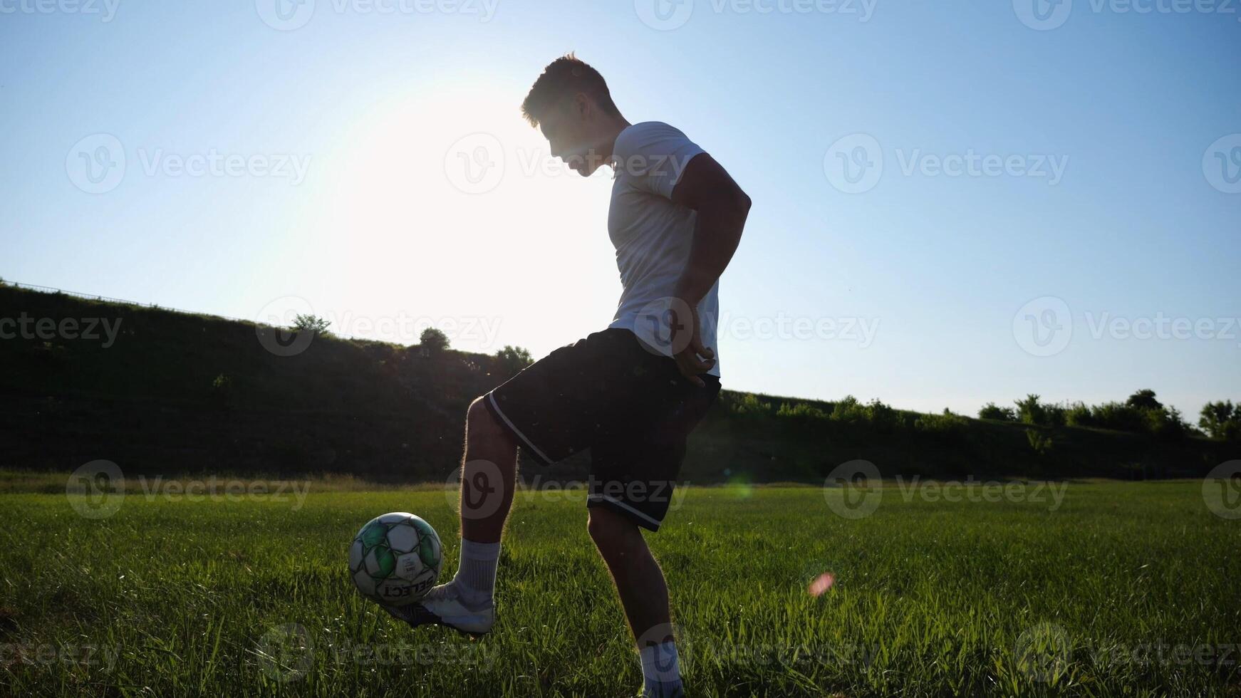 Sportsman juggling soccer ball on stadium at sunny day. Young man kicking ball at green field. Professional footballer practicing tricks at meadow with sunlight at background. Freestyle football. photo
