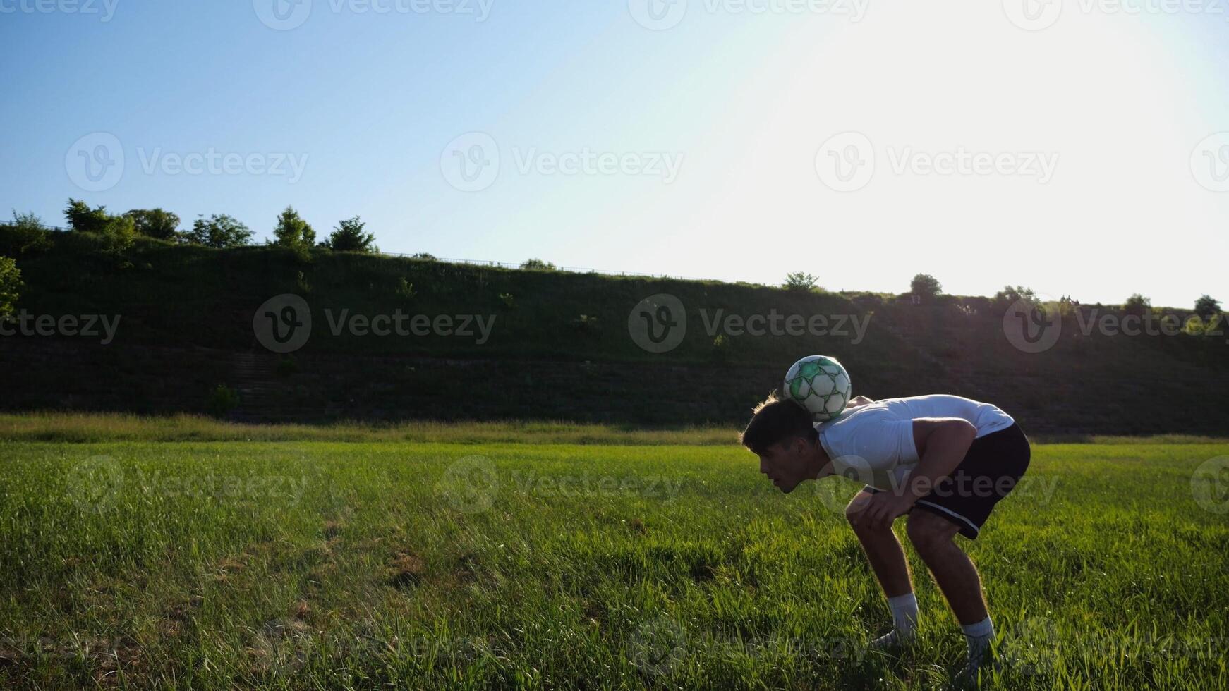 Sportsman juggling soccer ball on stadium at sunny day. Young man kicking ball at green field. Professional footballer practicing tricks at meadow with sunlight at background. Freestyle football. photo