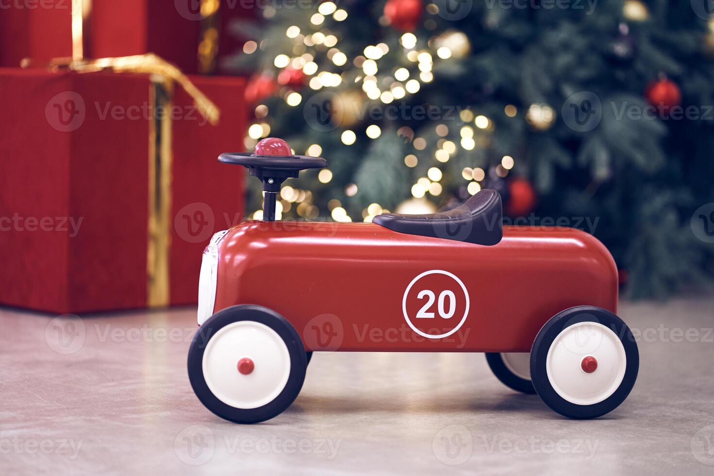 View of red children's retro car on the background of gifts in red boxes with golden bows, Christmas tree decorated garland, string light, red and blue balls. Background, copy space, bokeh photo