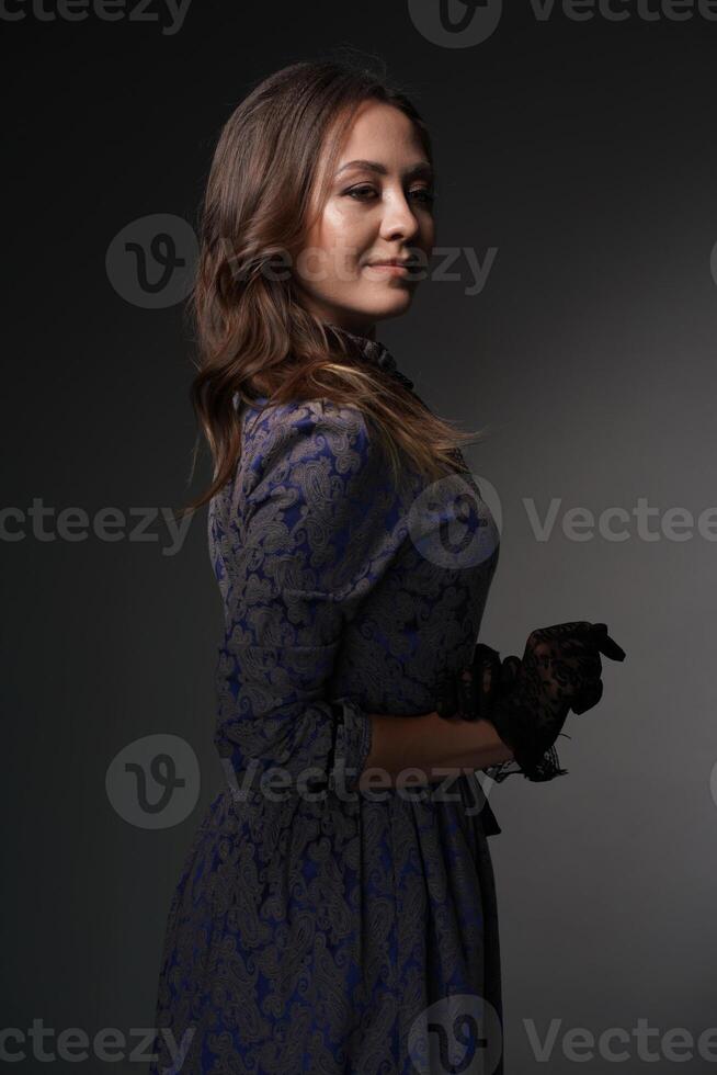 A woman stands with her back to the camera, looking over her shoulder in a blue retro dress in the style of the 1900s. A woman with loose hair. Historical fashion of the 19th century photo