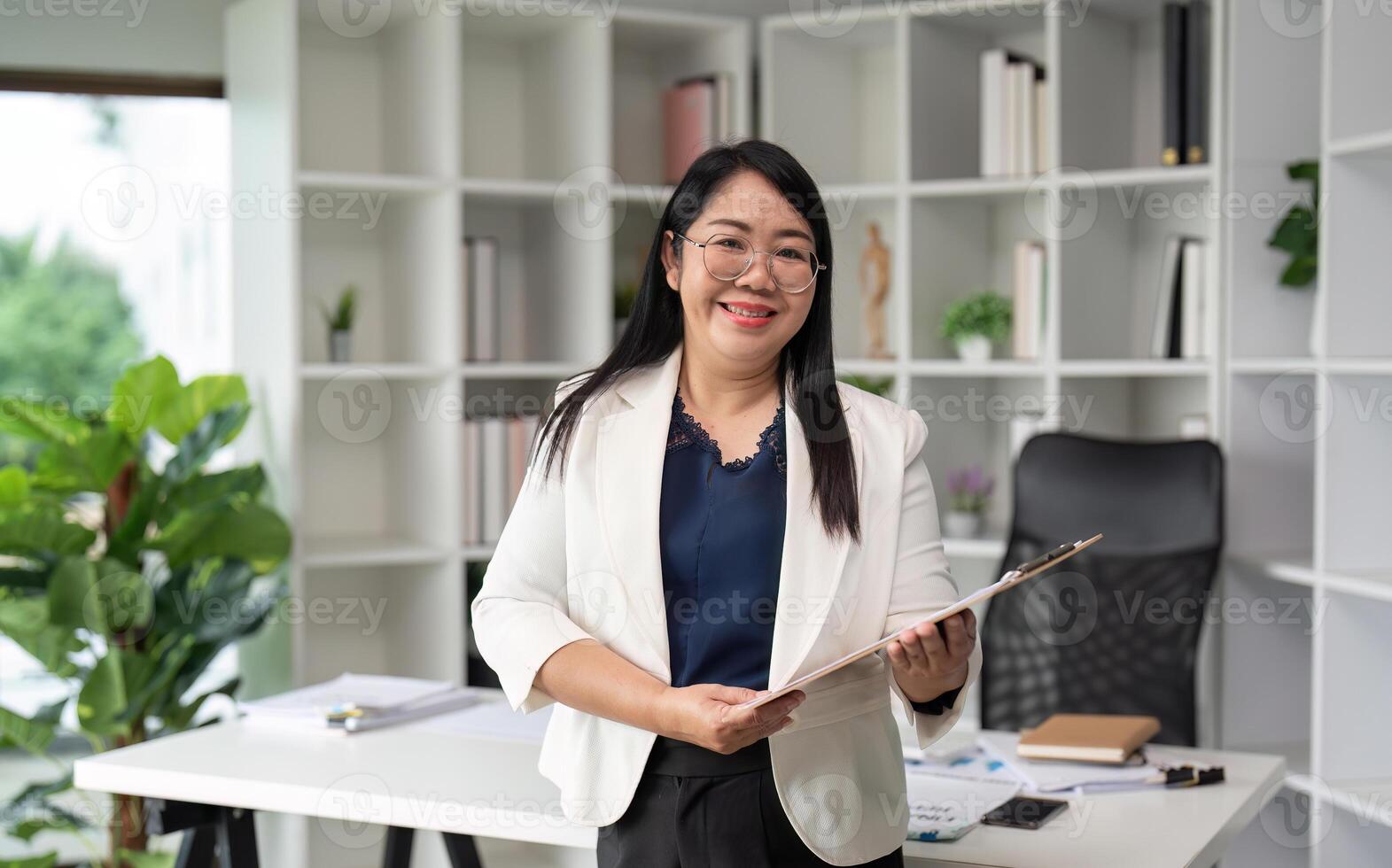 Middle-aged Business Woman Smiling While Doing Paperwork in a Modern Office Setting with Shelves and Green Plants photo