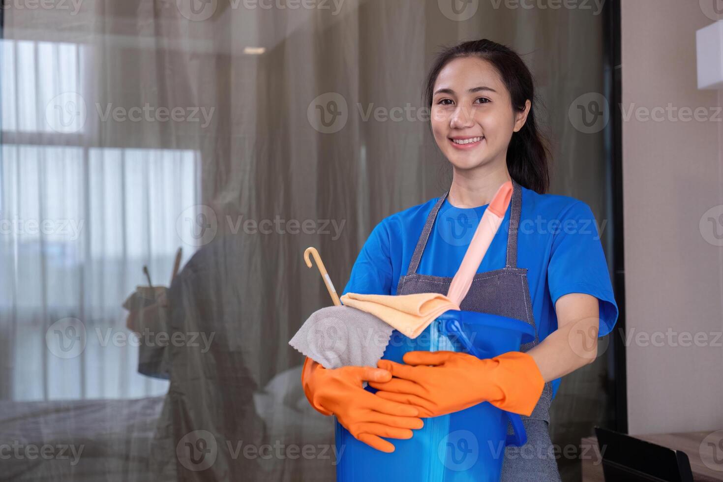 Professional Maid Cleaning Modern Room with Cleaning Supplies, Wearing Uniform and Gloves, Smiling at Camera photo
