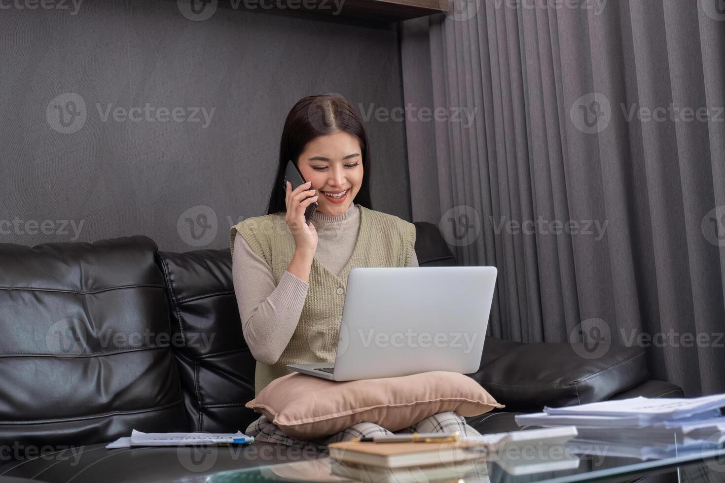 Young Woman Working from Home on Laptop While Talking on Phone, Sitting on Couch in Modern Living Room photo