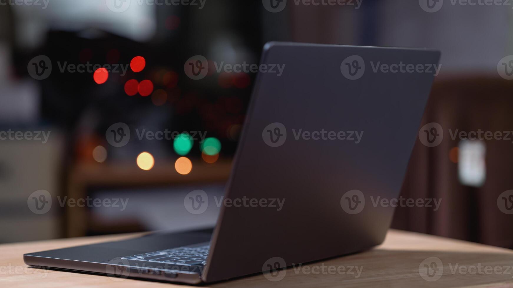 A laptop is open on a wooden table. The laptop is black and the screen is off. The table is in a dimly lit room photo