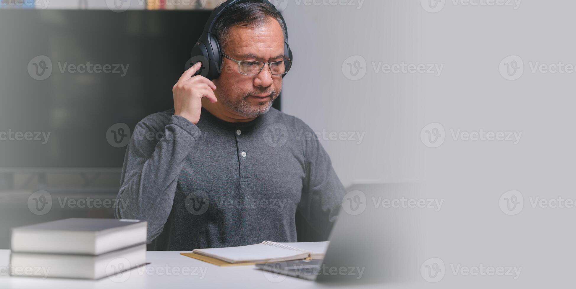 A man wearing headphones is sitting at a desk with a laptop and a stack of books. Adult Learning, Seniors in technology classes with headset. photo