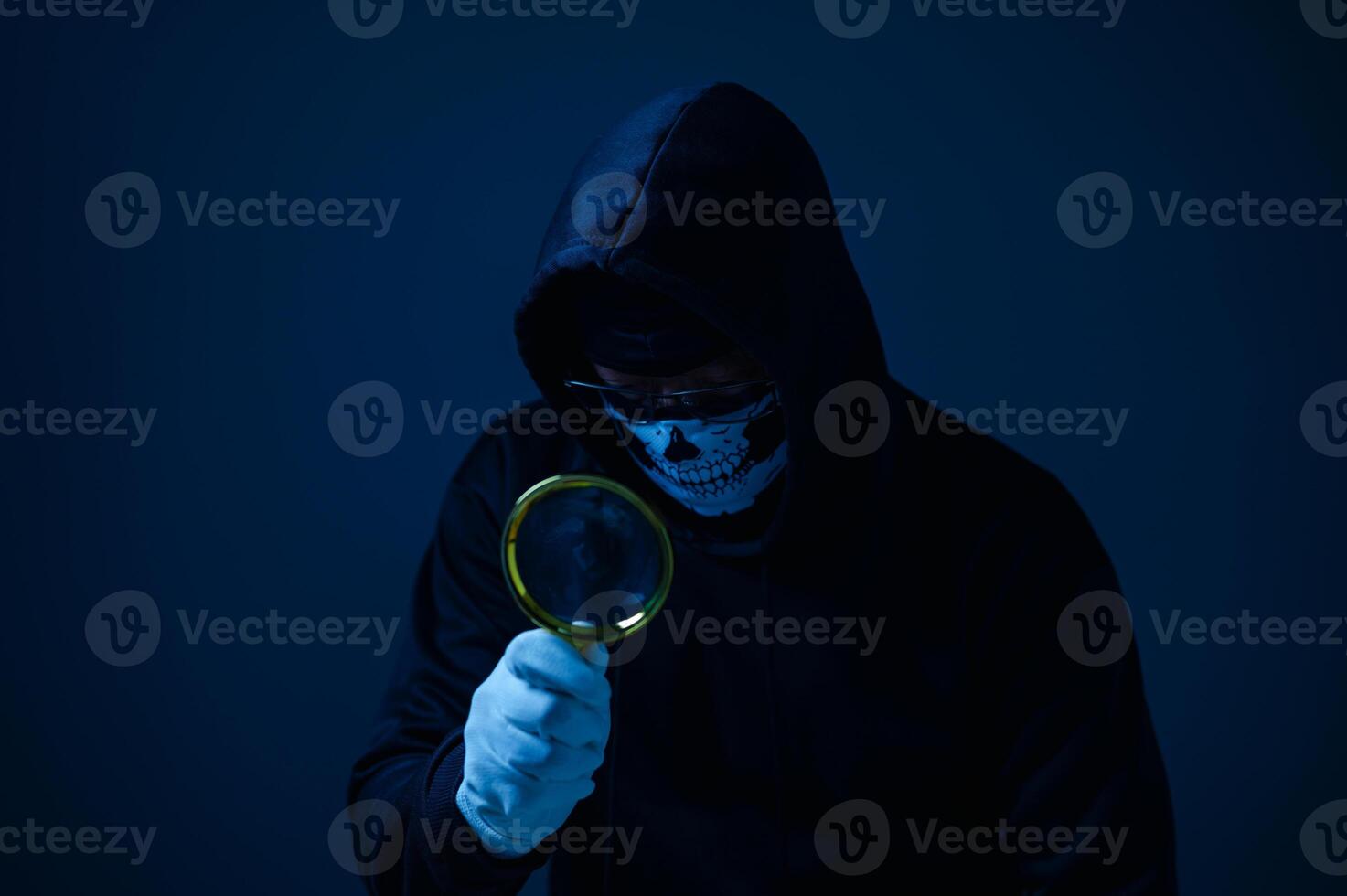 A hacker man in a hoodie is holding a magnifying glass. He is looking at something, but it is not clear what he is looking at. The image has a mysterious and suspenseful mood, as the man's actions photo