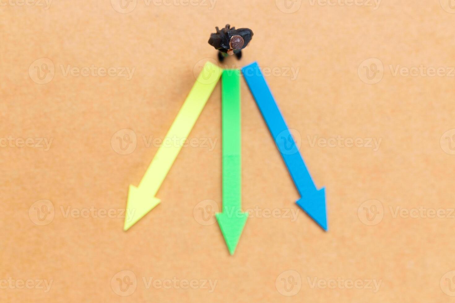A miniature people stands at the intersection of colorful arrows on a light brown background photo