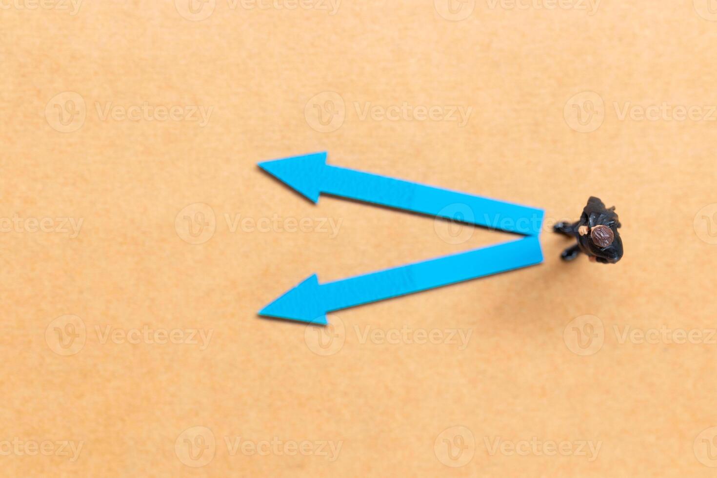 A miniature people stands at the intersection of colorful arrows on a light brown background photo