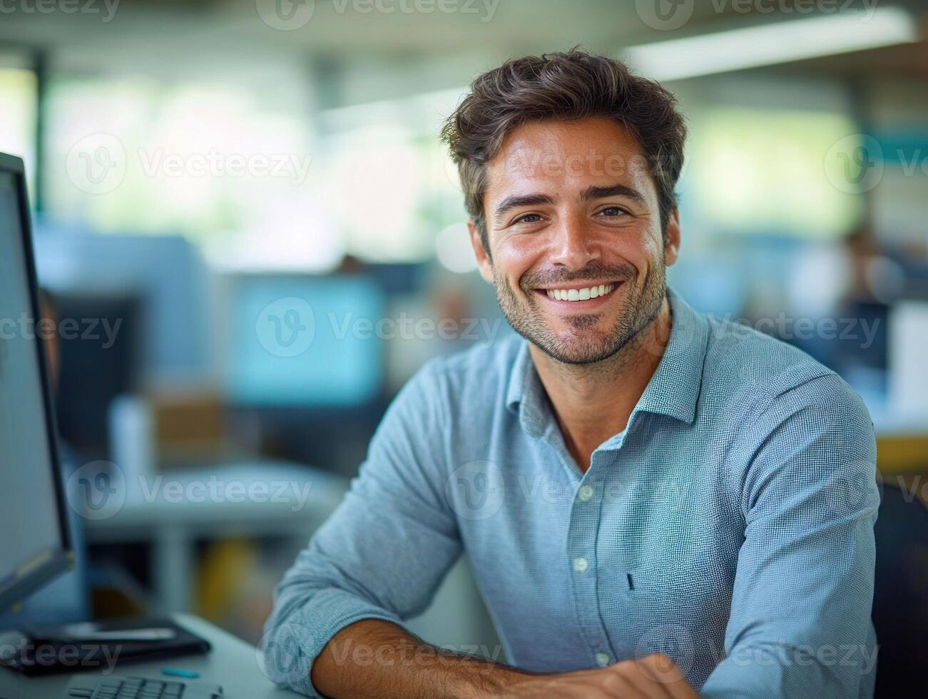 A man smiles confidently at his desk, surrounded by a bright, collaborative office space photo