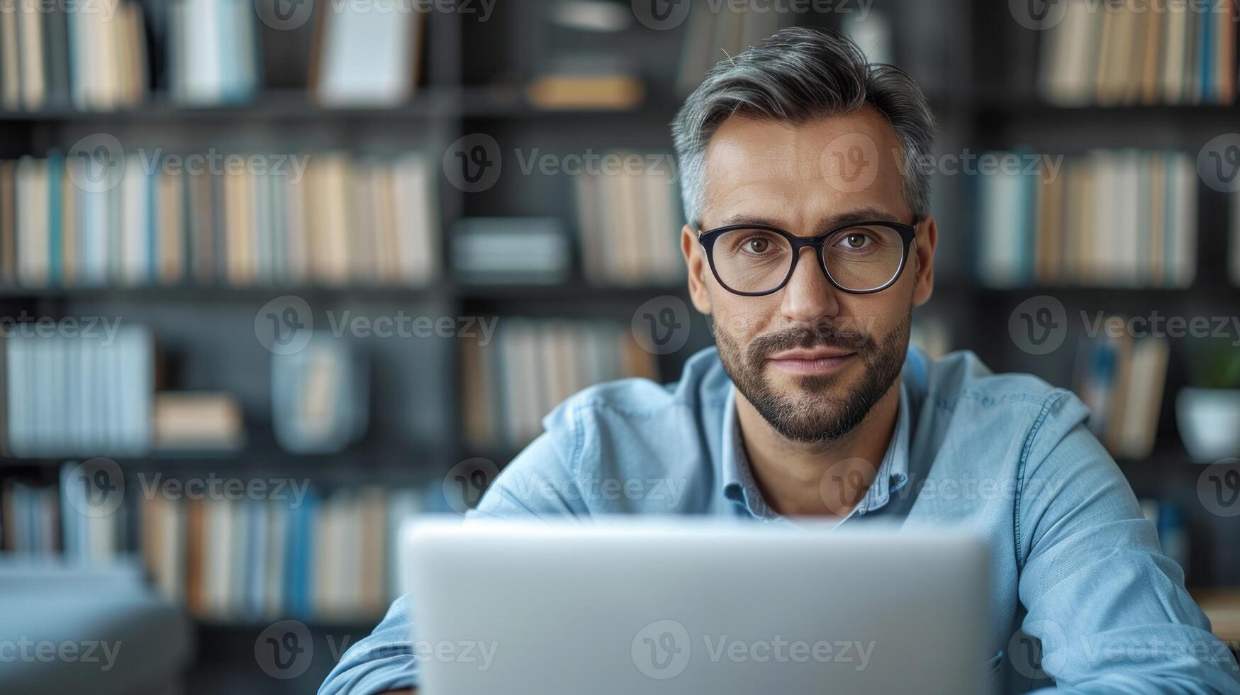 Man with graying hair wearing glasses looks directly at camera while working on laptop in an office photo