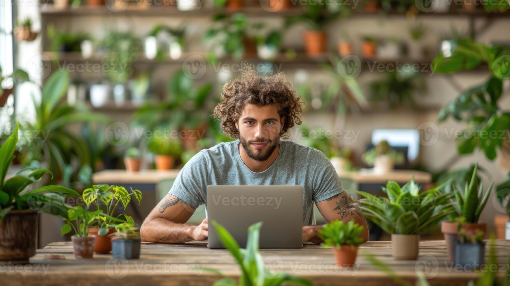 A man sits at a wooden table, working on a laptop, surrounded by various potted plants photo