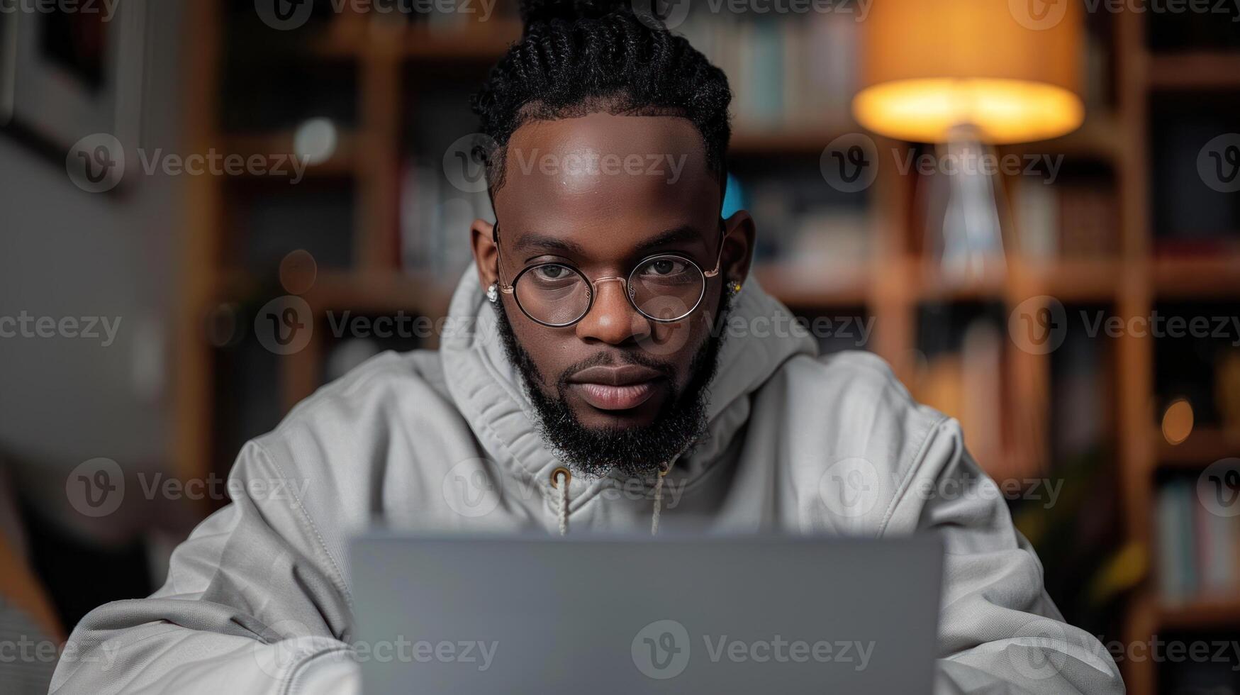 A man with dreadlocks wearing glasses sits at a desk in front of a laptop, looking intently at the screen photo