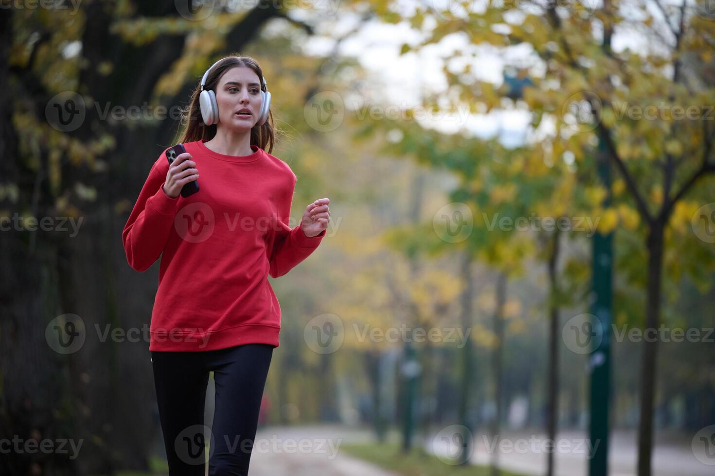 Young beautiful woman running in autumn park and listening to music with headphones on smartphone photo