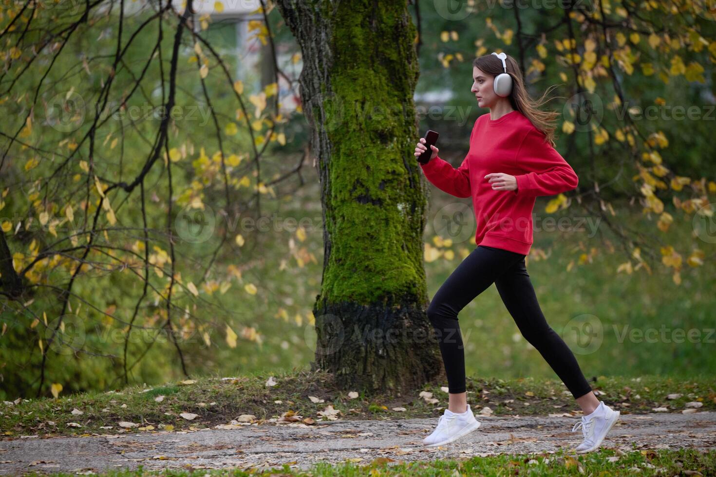 Young beautiful woman running in autumn park and listening to music with headphones on smartphone photo