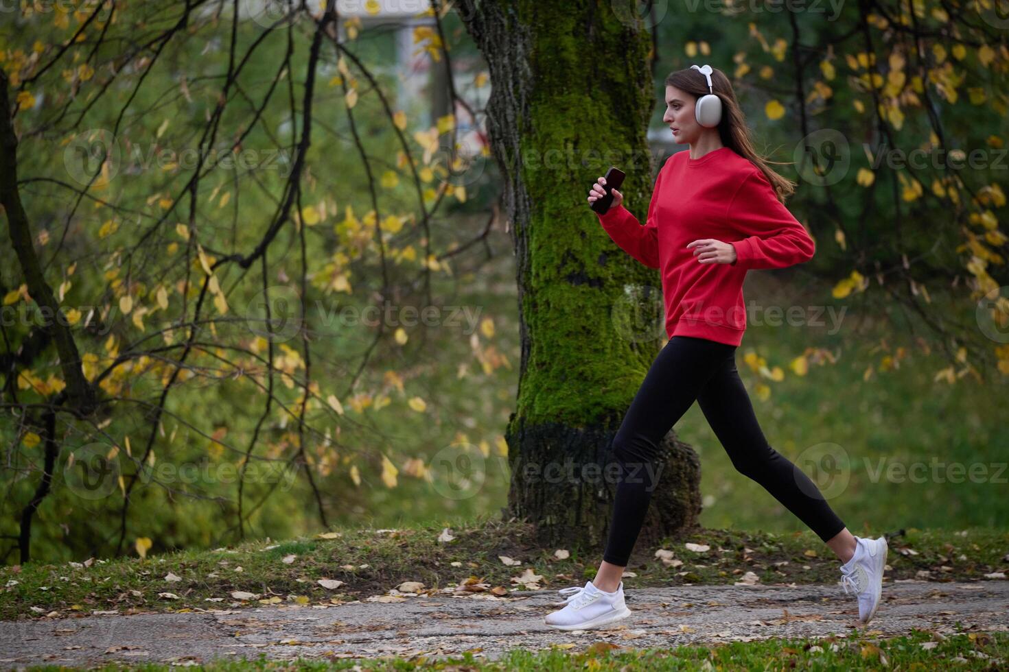 Young beautiful woman running in autumn park and listening to music with headphones on smartphone photo