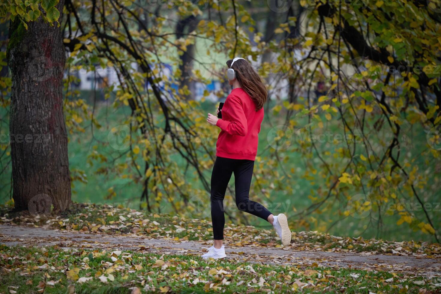 Young beautiful woman running in autumn park and listening to music with headphones on smartphone photo
