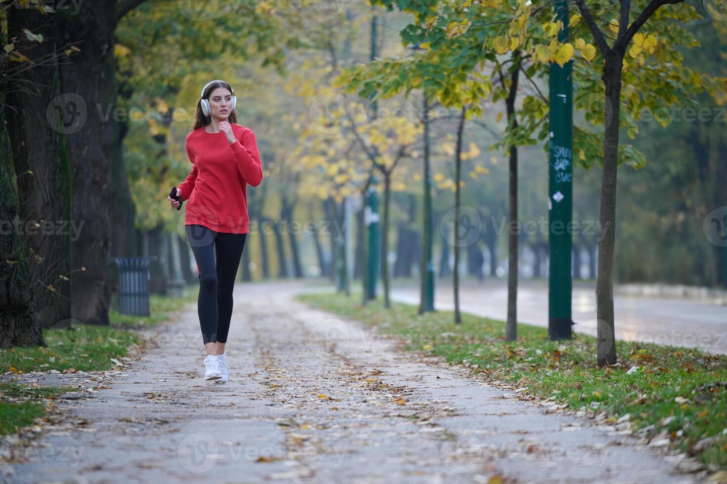 Young beautiful woman running in autumn park and listening to music with headphones on smartphone photo