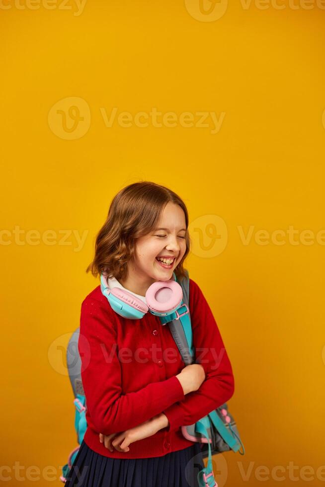 Smiling Teenage Schoolgirl With Backpack and Headphones Against Yellow Background photo
