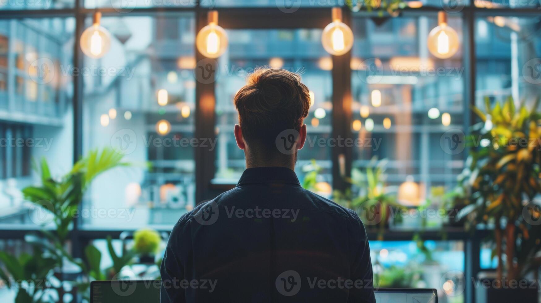 A man stands in a modern office filled with plants and warm hanging lights, looking out a large window at an urban environment. photo