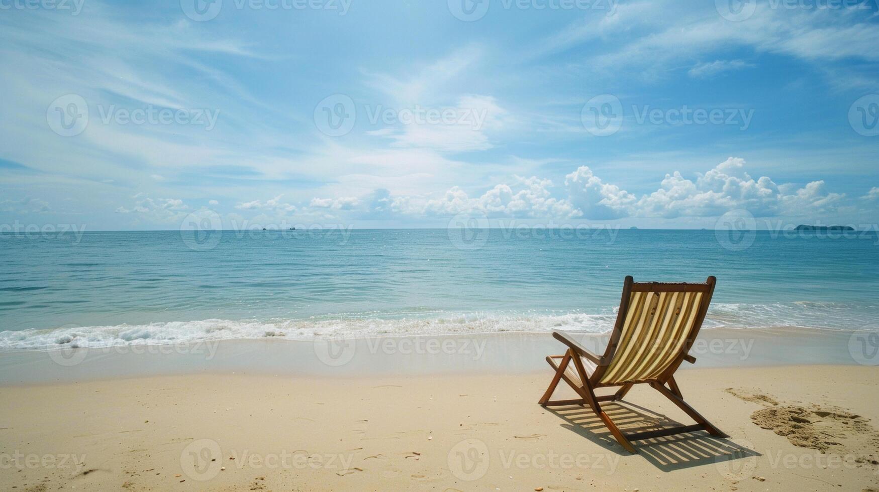 An empty deck chair facing the calm ocean on a serene sandy beach under a bright blue sky with scattered clouds. photo