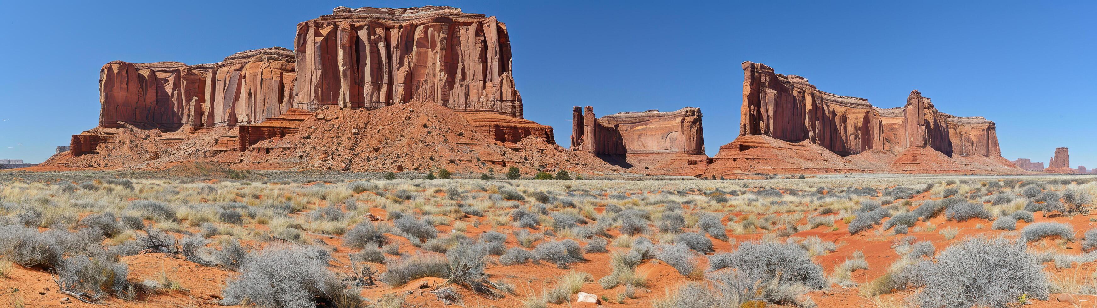 A panoramic photo captures red rock formations towering over a valley with a clear blue sky and lush green shrubs