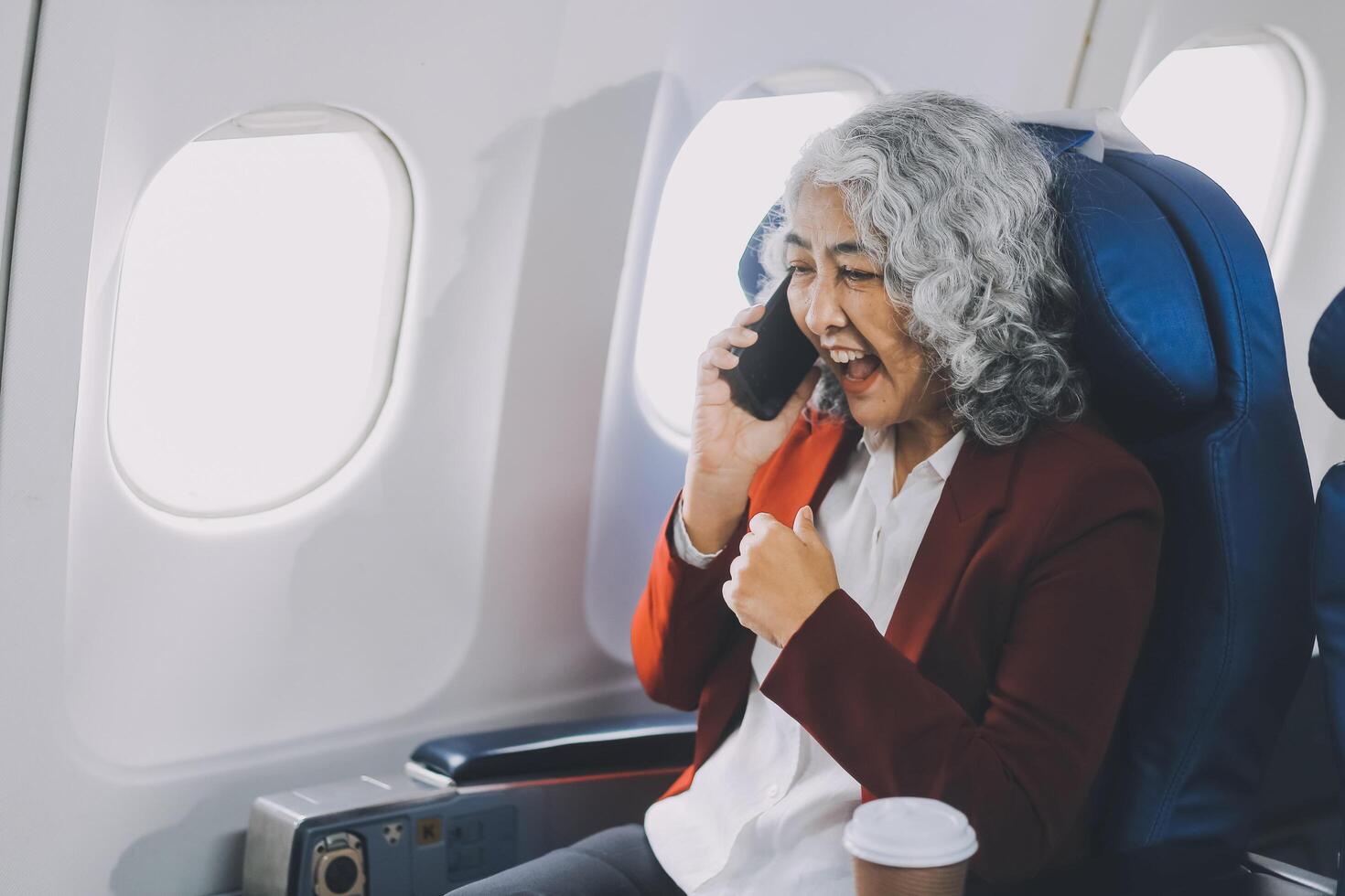 Portrait of a successful Asian businesswoman or entrepreneur in a formal suit on an airplane sitting in business class using a phone, computer laptop. Travel in style, work with grace. photo