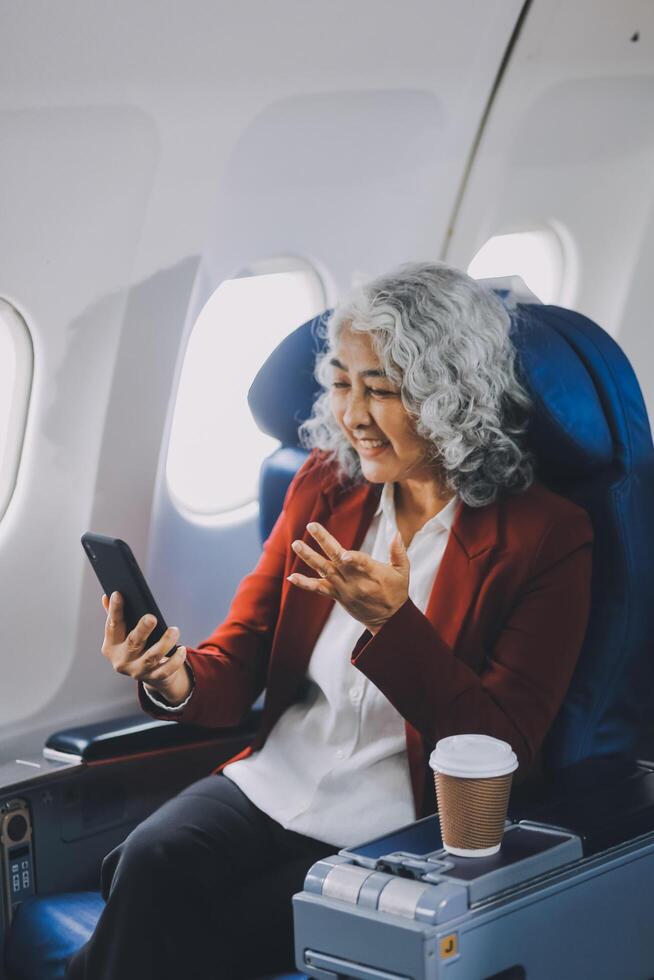 Portrait of a successful Asian businesswoman or entrepreneur in a formal suit on an airplane sitting in business class using a phone, computer laptop. Travel in style, work with grace. photo