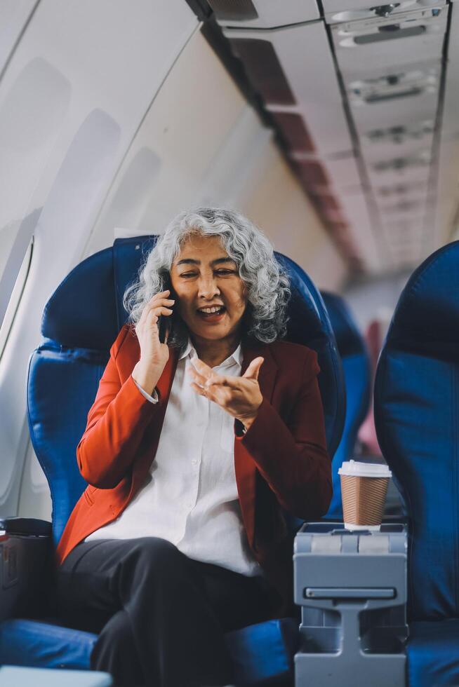 Portrait of a successful Asian businesswoman or entrepreneur in a formal suit on an airplane sitting in business class using a phone, computer laptop. Travel in style, work with grace. photo