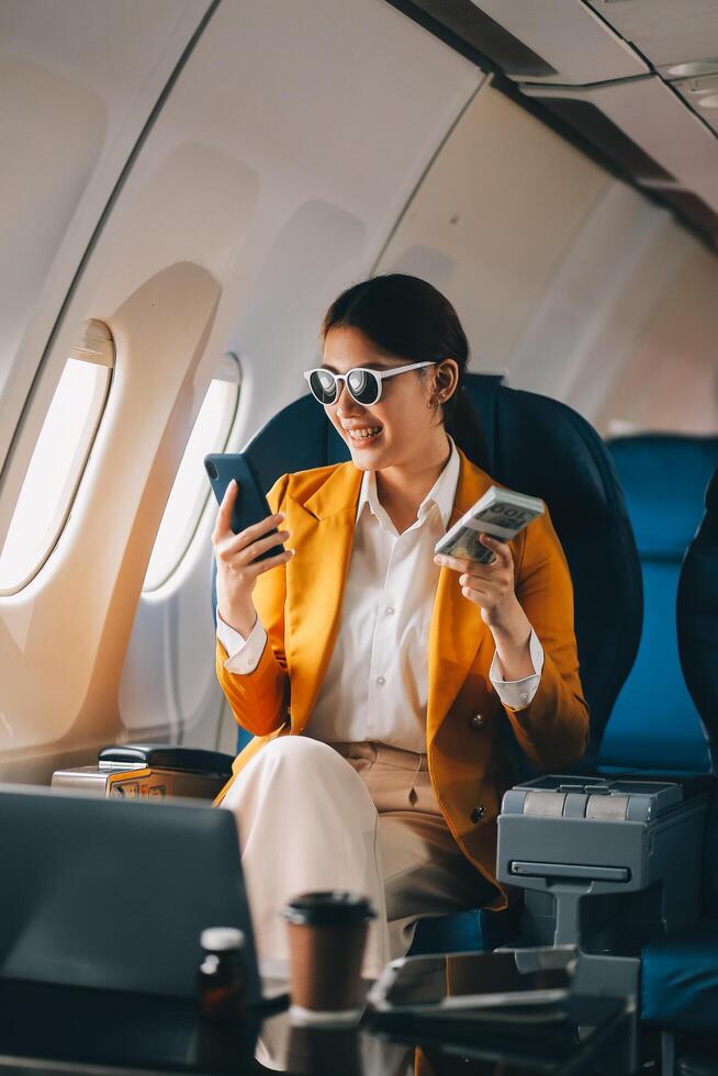 A young Asian woman, seated as an airplane passenger, contemplates dollar bills, reflecting on lucrative opportunities and aspirations towards billionaire status. photo