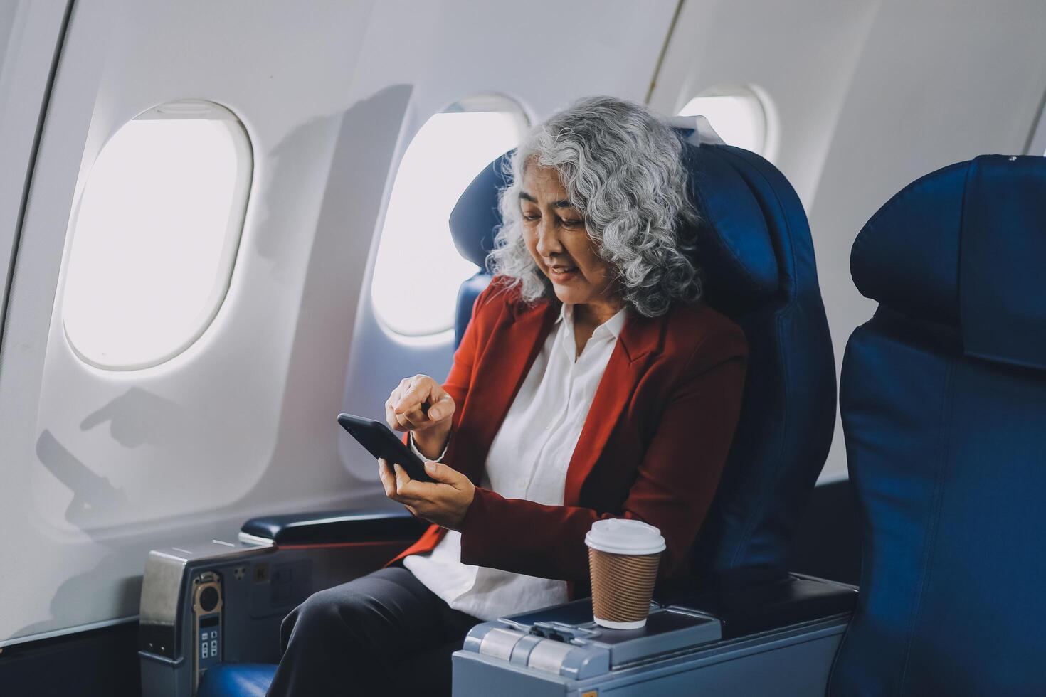 Portrait of a successful Asian businesswoman or entrepreneur in a formal suit on an airplane sitting in business class using a phone, computer laptop. Travel in style, work with grace. photo