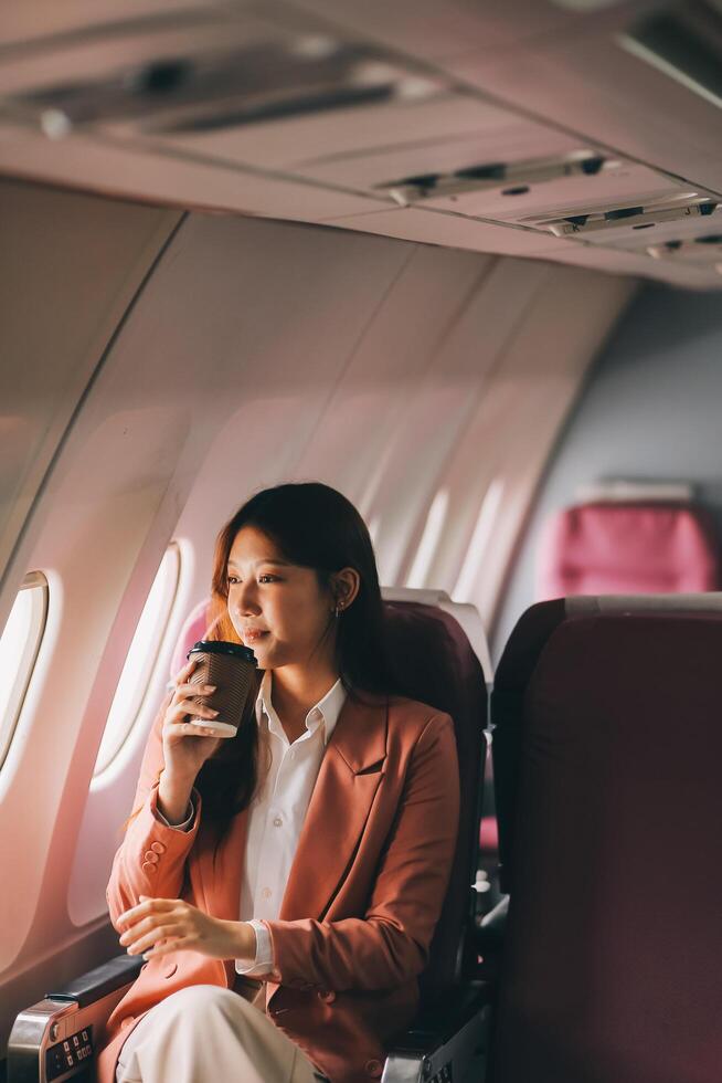 Portrait of a successful Asian businesswoman or entrepreneur in a formal suit on an airplane sitting in business class using a phone, computer laptop. Travel in style, work with grace. photo