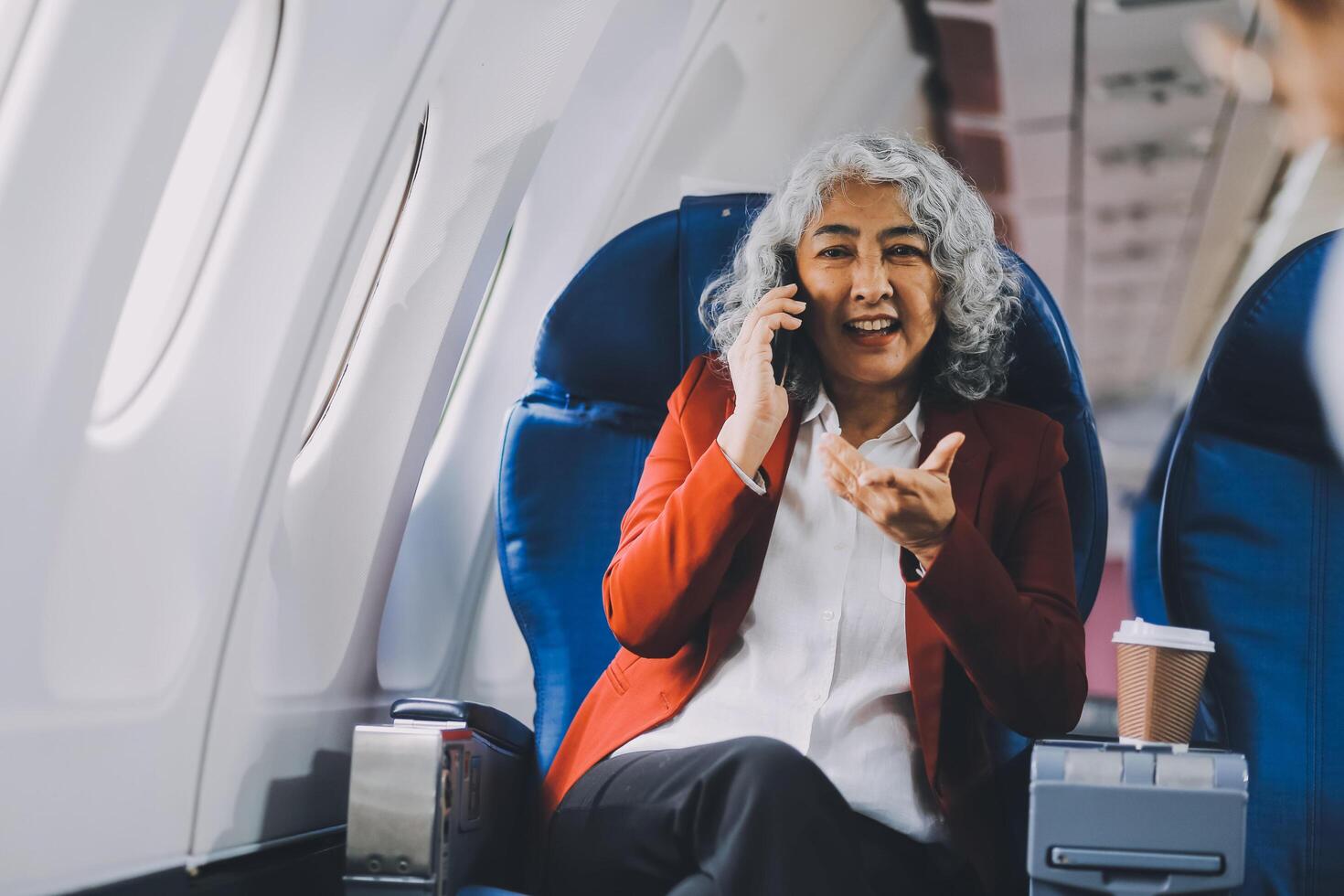 Portrait of a successful Asian businesswoman or entrepreneur in a formal suit on an airplane sitting in business class using a phone, computer laptop. Travel in style, work with grace. photo