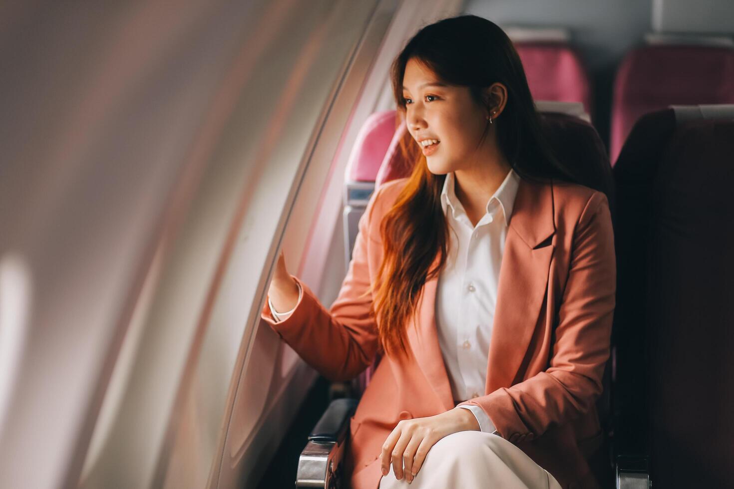 Portrait of a successful Asian businesswoman or entrepreneur in a formal suit on an airplane sitting in business class using a phone, computer laptop. Travel in style, work with grace. photo