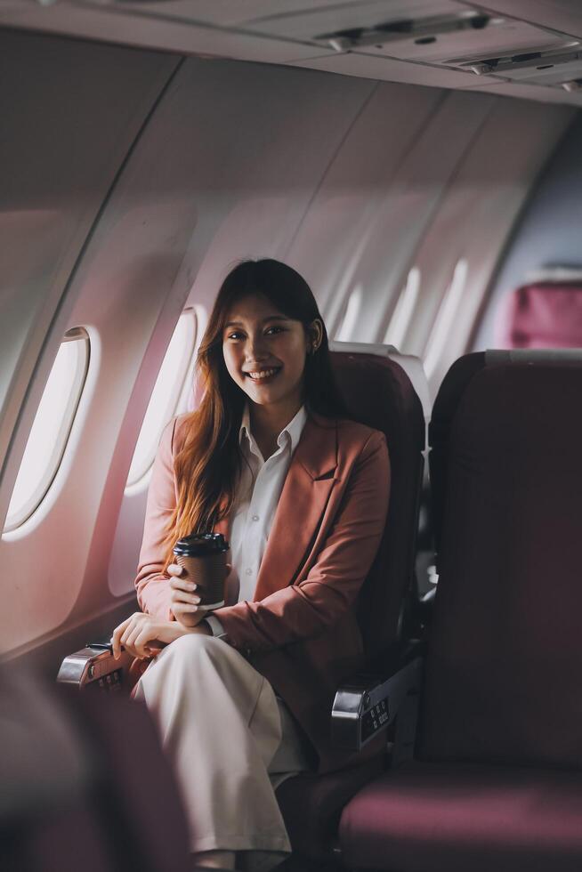 Portrait of a successful Asian businesswoman or entrepreneur in a formal suit on an airplane sitting in business class using a phone, computer laptop. Travel in style, work with grace. photo