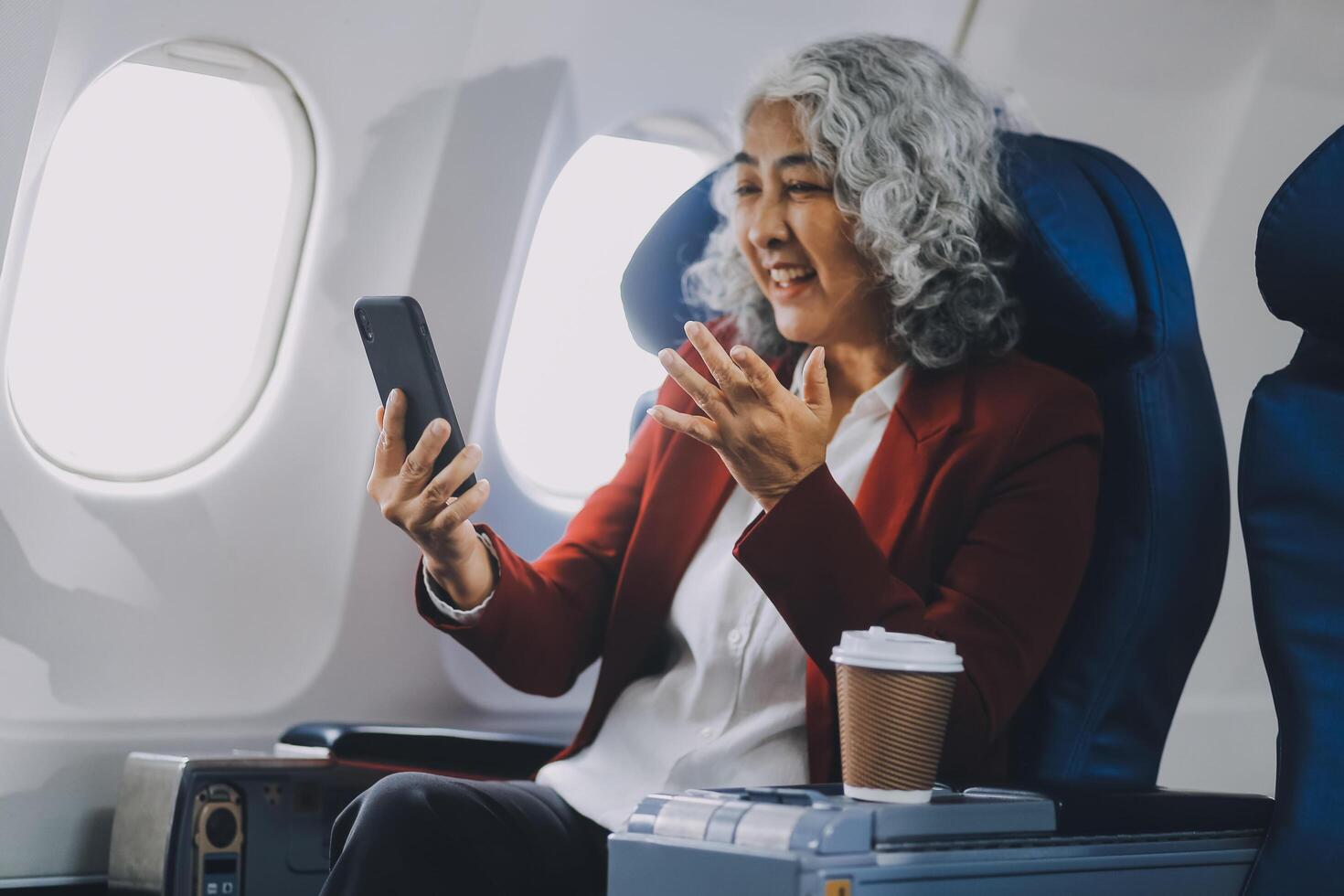 Portrait of a successful Asian businesswoman or entrepreneur in a formal suit on an airplane sitting in business class using a phone, computer laptop. Travel in style, work with grace. photo