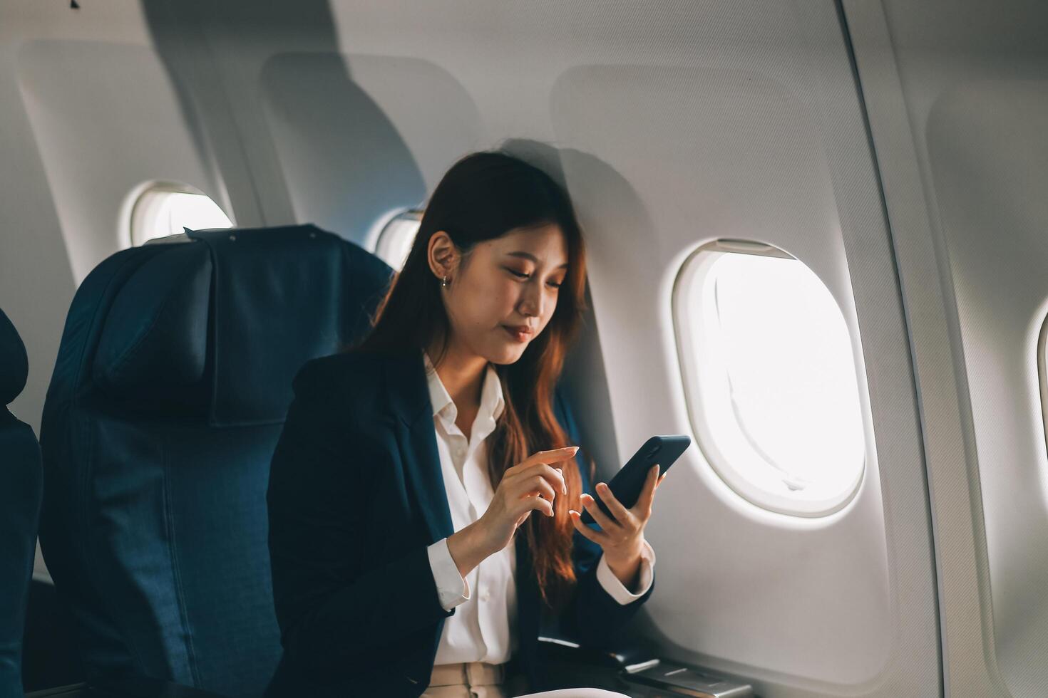 A young Asian female airplane passenger sits by the window during the flight, holding her boarding pass, with her carry-on luggage stored overhead, wearing her seatbelt, and enjoying the journey. photo